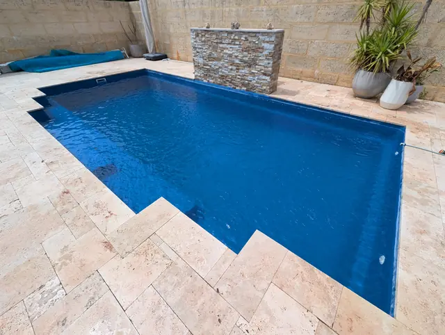 Deep blue pool with travertine paving and a stone water feature